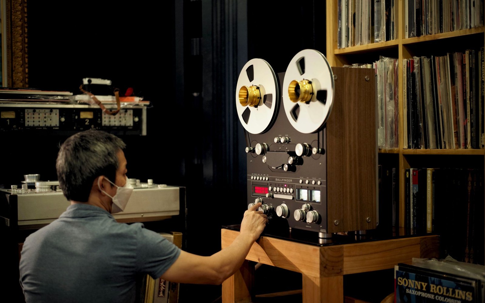 A man adjusts controls on a reel-to-reel tape machine beside shelves of vinyl records