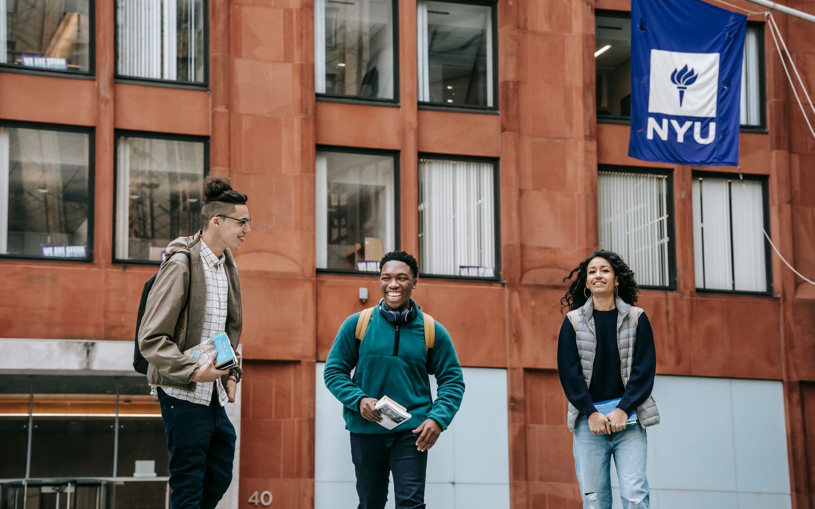 Three students chat and laugh outside a brick building with an NYU banner hanging above