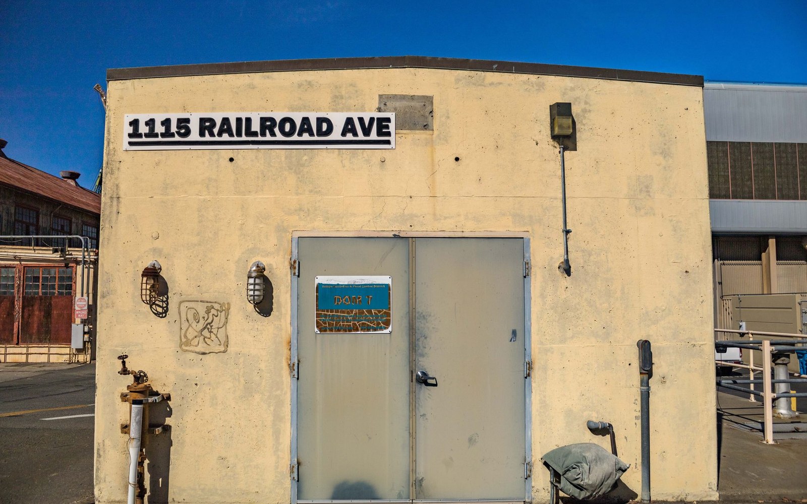 A tan concrete building marked "1115 Railroad Ave" with a metal door and wall lamps