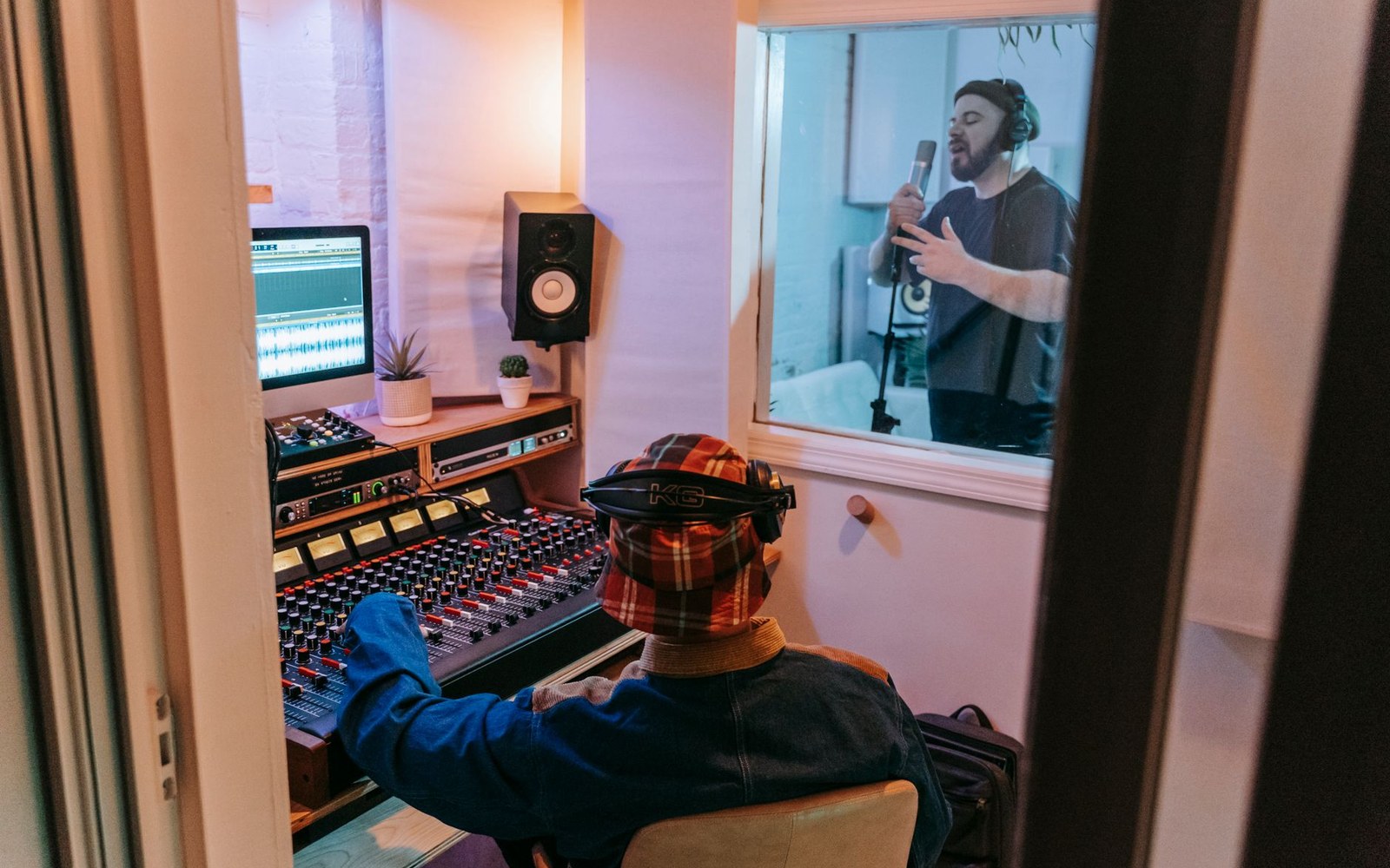 Adult musicians and a recording engineer in a wide studio room, all visible in one frame, shot from the back of the room toward the control-room glass